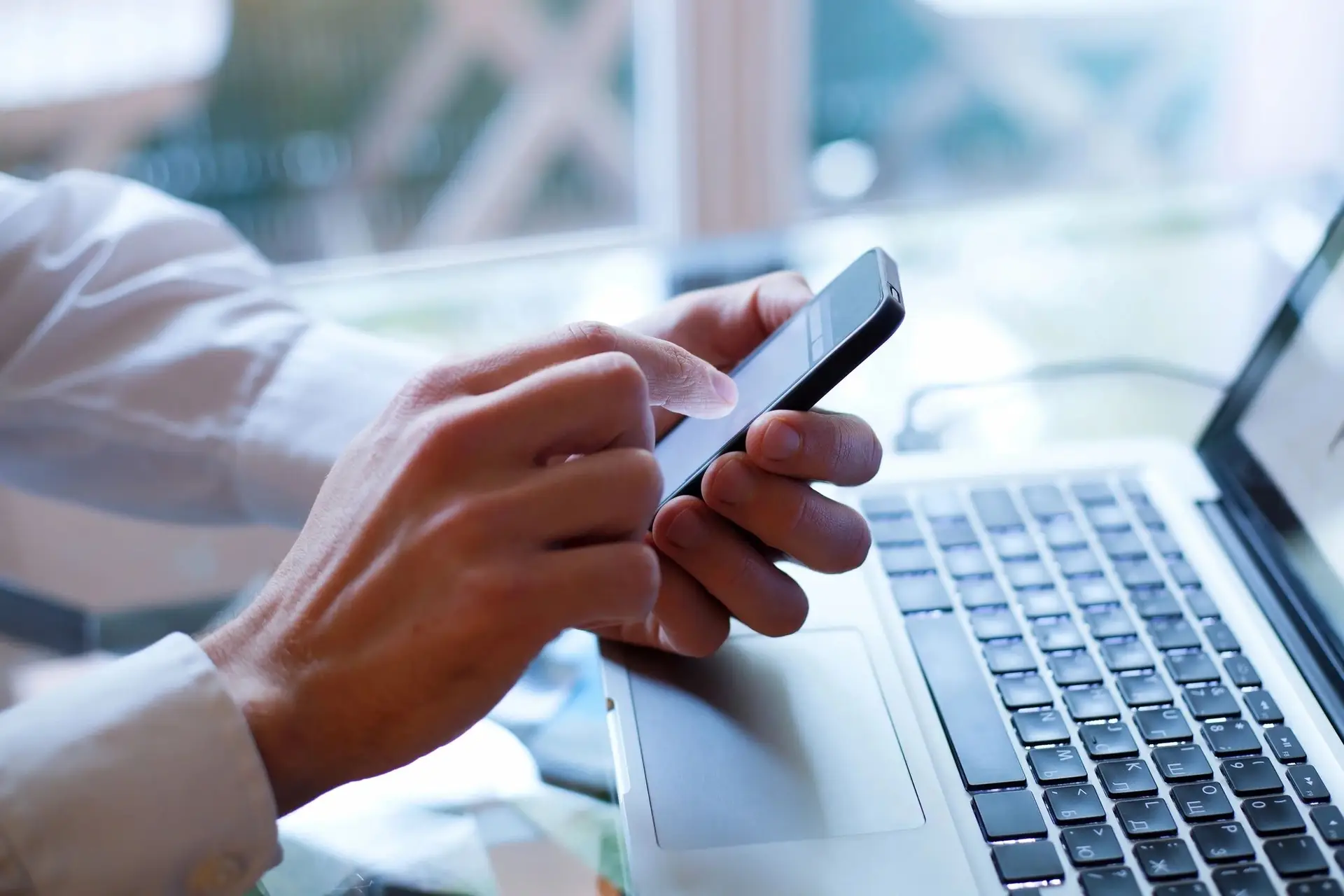 Person using a smartphone while resting hands over a laptop keyboard, indoors near a window.