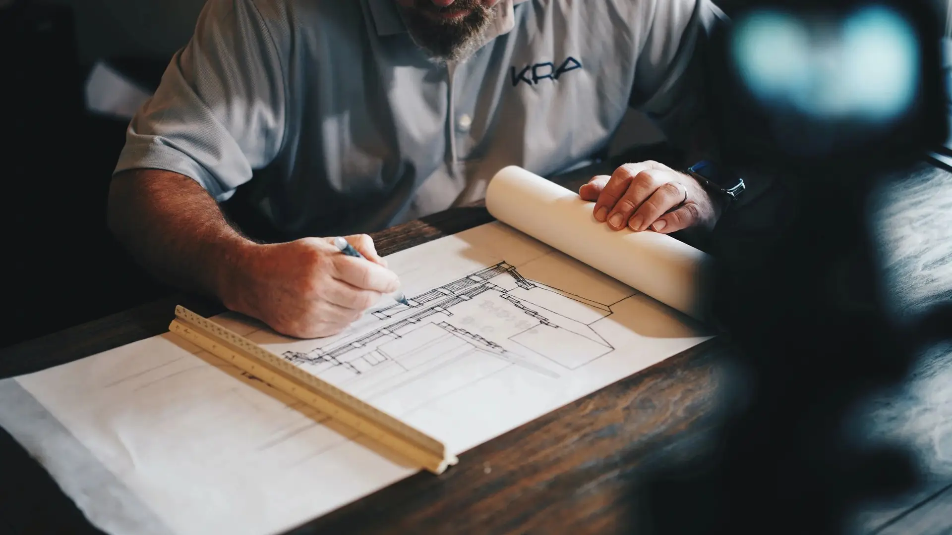 qtq80-2rn1NY Man drawing architectural plans on blueprints with a pencil, a ruler resting nearby on a wooden table